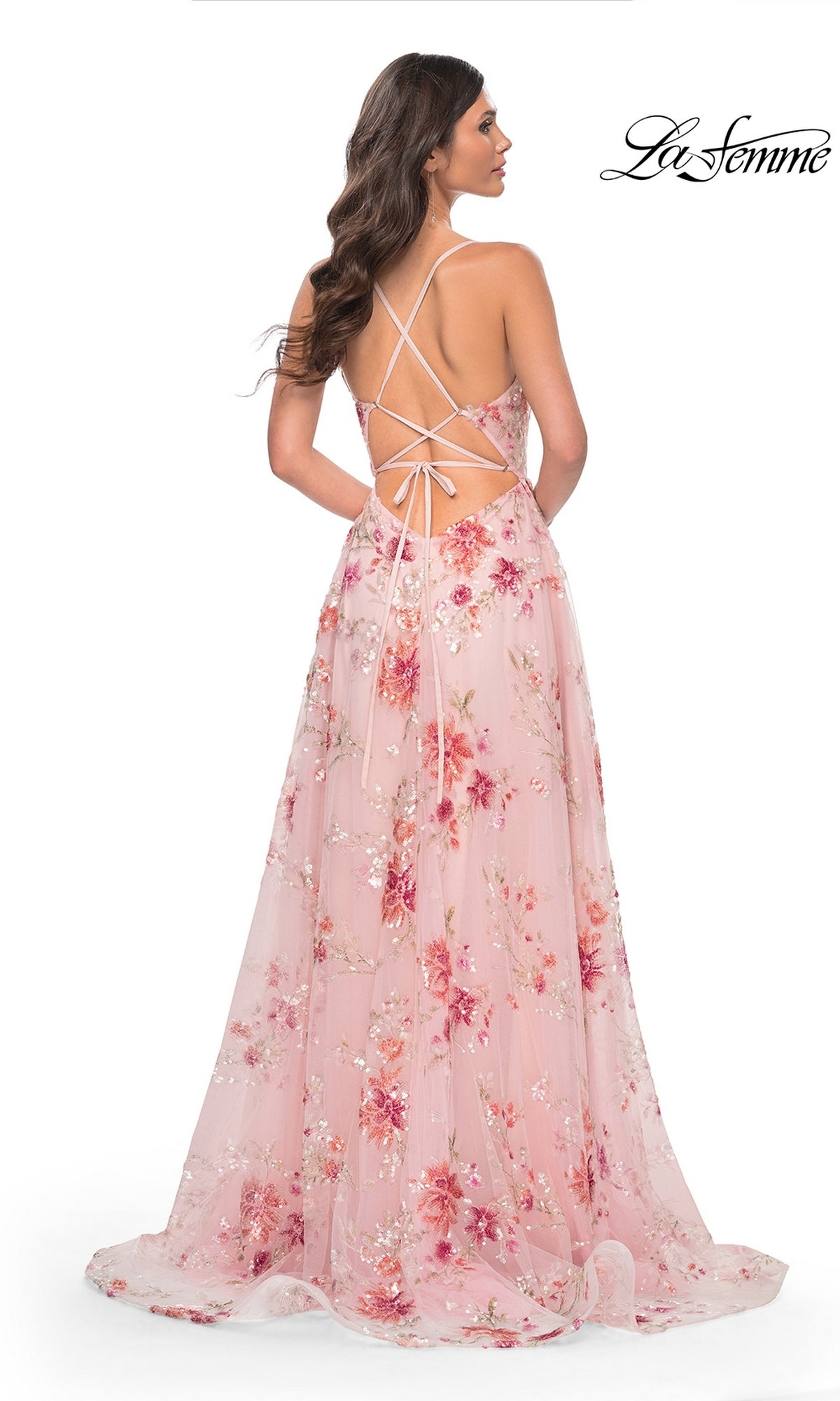 A woman in the La Femme Floral-Sequin Long A-Line Prom Dress 32031 stands turned away, showcasing its floor-length, backless design with crisscross straps and a flowing, semi-sheer pink floral skirt for an elegant look.